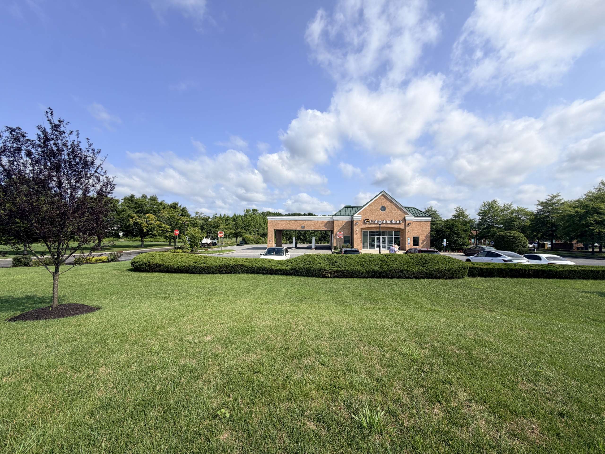 Front view of a brick Columbia Bank building with a green roof, surrounded by trees, in a parking lot under a blue sky with scattered clouds.