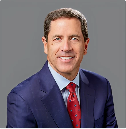 Professional headshot of a smiling man in a navy suit and red tie against a gray backdrop.